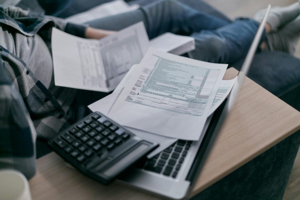 A young adult using a calculator and laptop to manage finances at home, surrounded by papers.