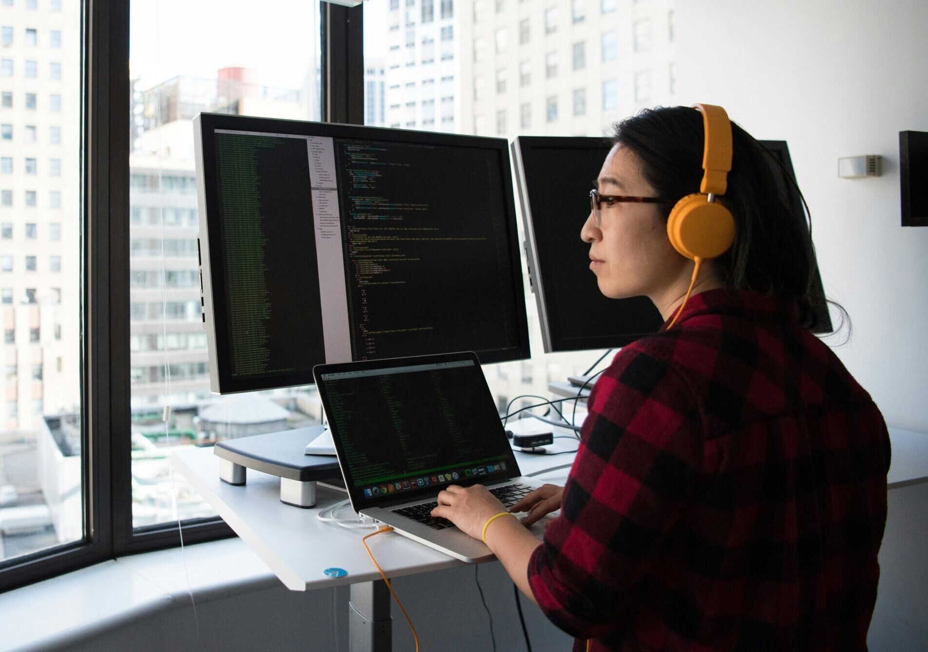 A businesswoman coding at a standing desk with city views through large windows, wearing headphones.