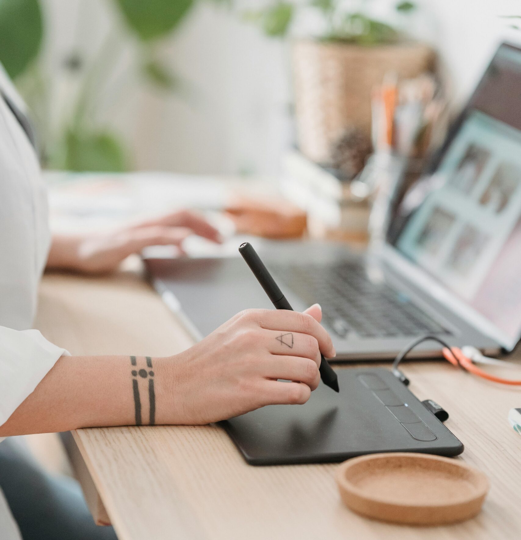 Crop anonymous female browsing netbook and working on graphic tablet with stylus while sitting at desk