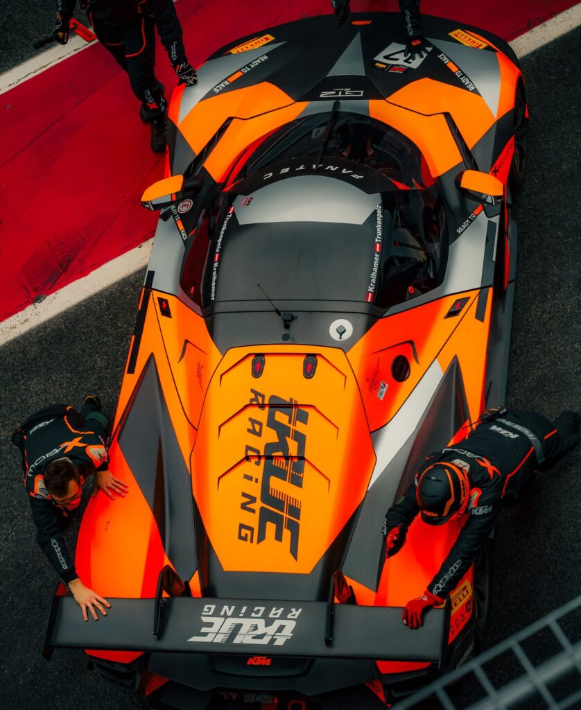Bright orange race car with pit crew, captured at CT, España. High-speed, dynamic scene.